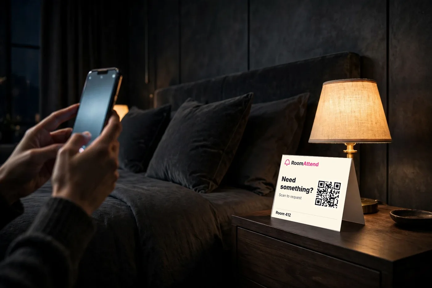 Guest in a dark hotel room holding a phone beside the bed, with a RoomAttend QR card on the nightstand
